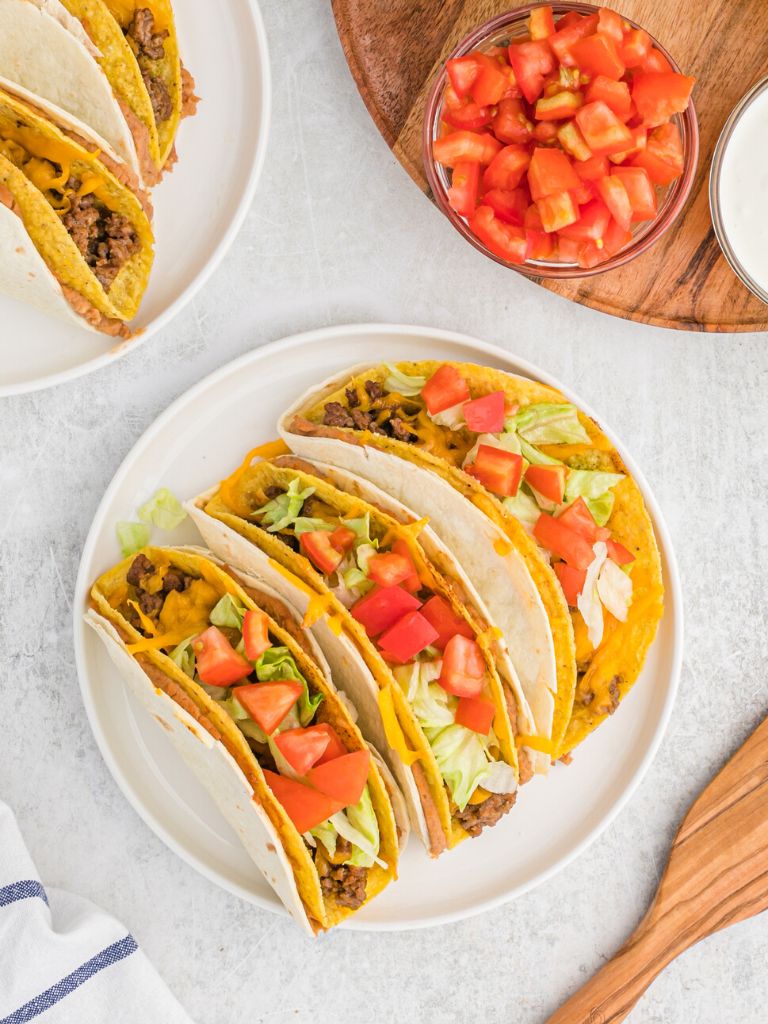 Overhead shot of tacos with a plate of them on the side and a bowl of tomatoes on top of a dark wooden cutting board. 