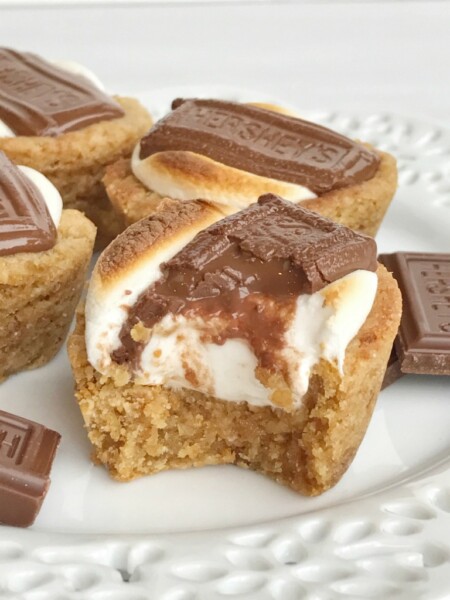 S'mores cookie cups are baked in a mini muffin pan. Graham cracker cookie base, with a toasted marshmallow, and a piece of gooey chocolate on top! Now you can enjoy campfire toasty s'more all year round for dessert.