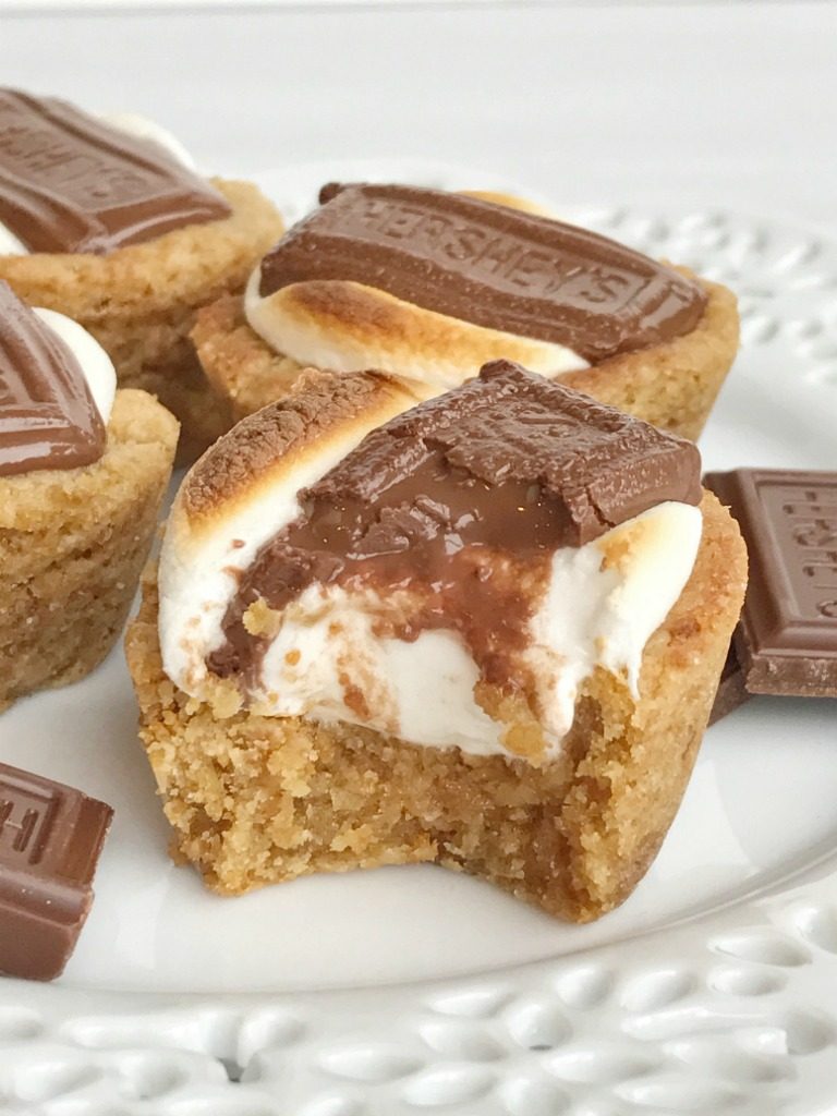 S'mores cookie cups are baked in a mini muffin pan. Graham cracker cookie base, with a toasted marshmallow, and a piece of gooey chocolate on top! Now you can enjoy campfire toasty s'more all year round for dessert. 