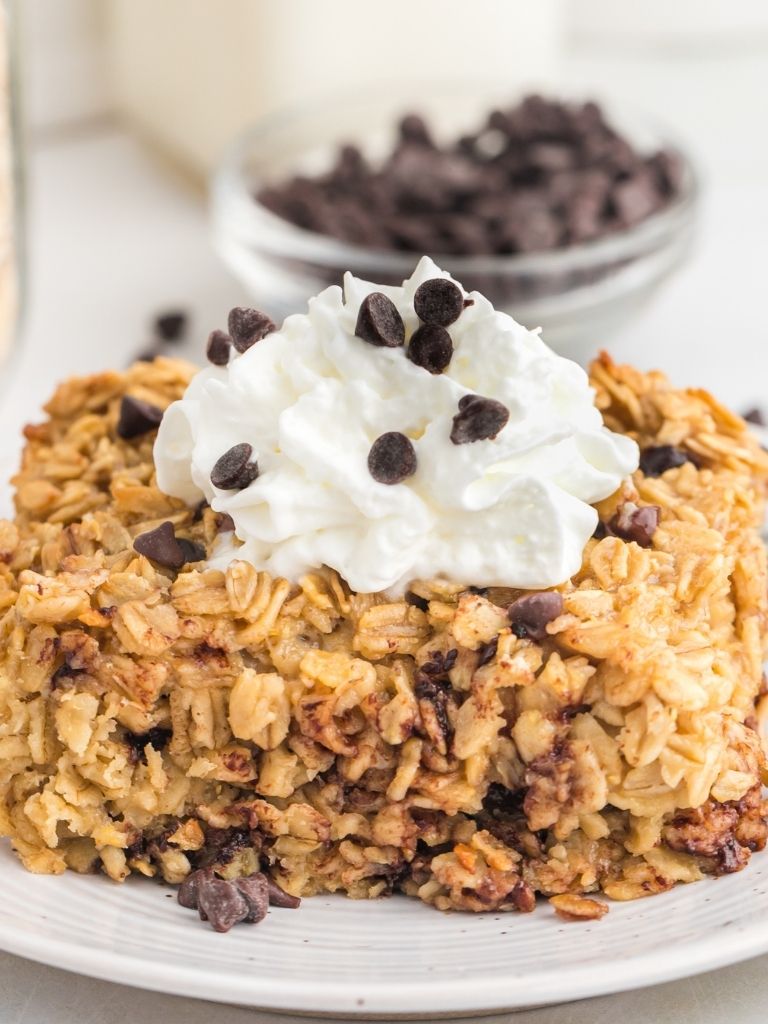 Square of baked oatmeal on a white plate topped with whipped cream and mini chocolate chips.