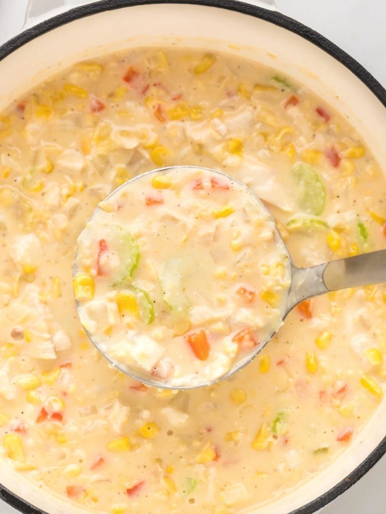 A white pot with chicken corn chowder inside of it and a soup ladle with some in it. 
