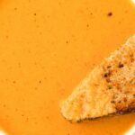 A white bowl with creamy tomato soup inside and a grilled cheese sandwich dipped into it.