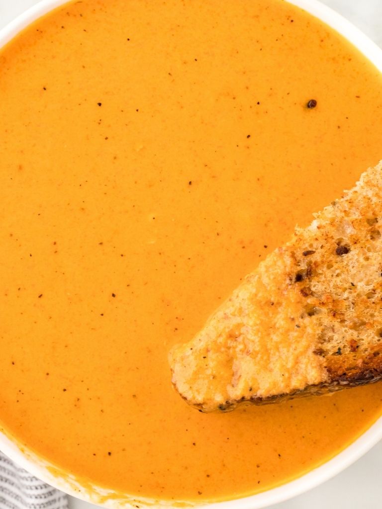 A white bowl with creamy tomato soup inside and a grilled cheese sandwich dipped into it.