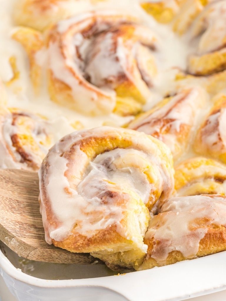 A wooden spatula picking up a cinnamon roll from a pan of them. 