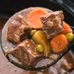 A scoop of apple cider beef stew with the slow cooker in the background.