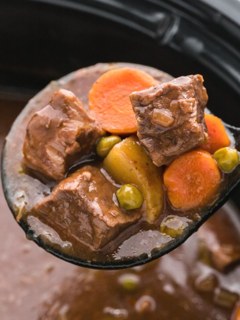 A scoop of apple cider beef stew with the slow cooker in the background.