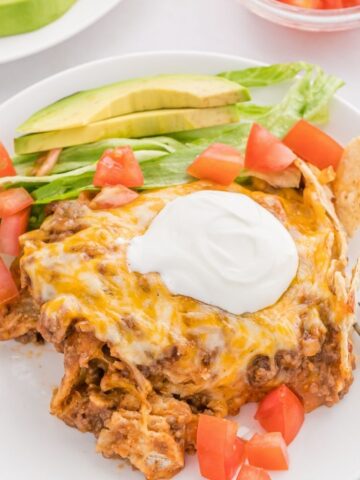 A serving of the casserole on a white plate topped with sour cream, tomatoes, lettuce, and avocado.