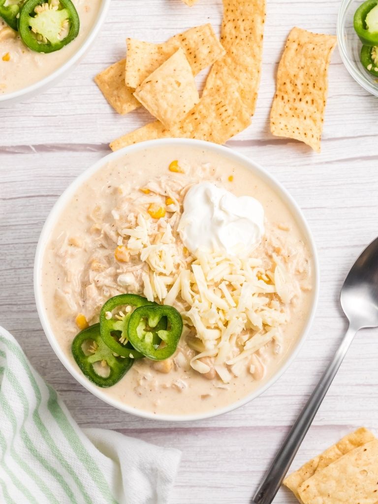 White chili inside a white bowl on a tables cape with chips and jalapeño slices. 