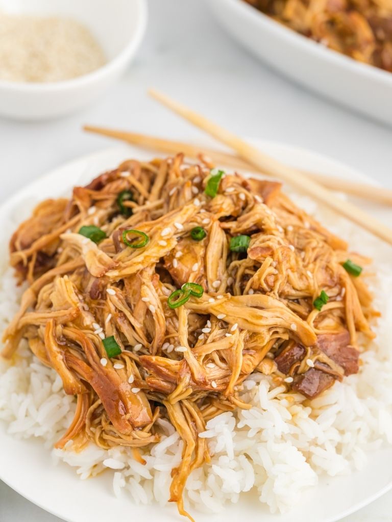 A white plate with shredded chicken in a sauce over white rice. Chopsticks in the background along with sesame seeds in a bowl.