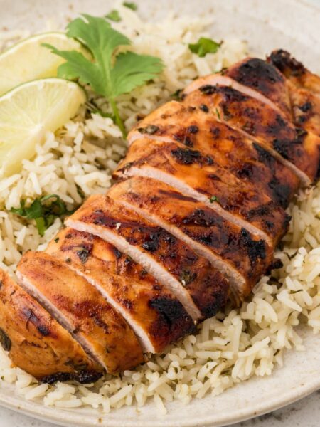 A piece of grilled chicken sliced sitting on top of a bed of rice on a white plate with lime wedges and cilantro leaves.