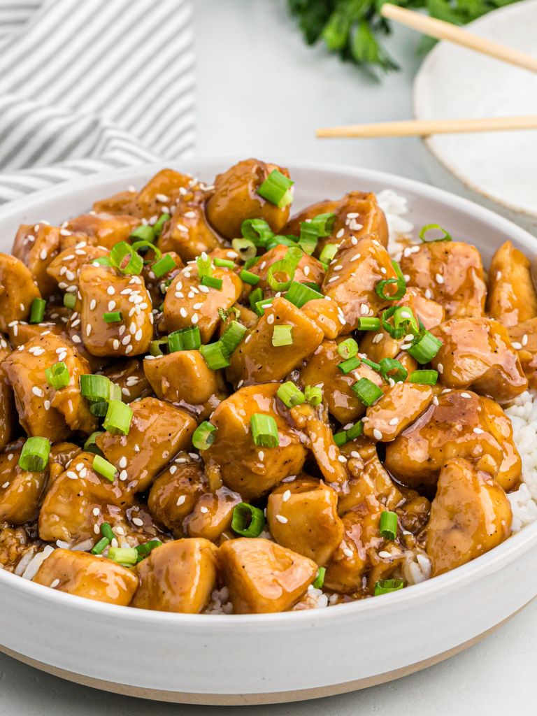 Bowl of teriyaki chicken topped with garnishes with a pair of chopsticks in the background of the photo. 