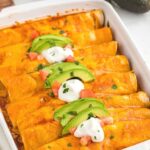 A white pan with enchiladas inside of it covered in red enchilada sauce and topped with avocado and sour cream.