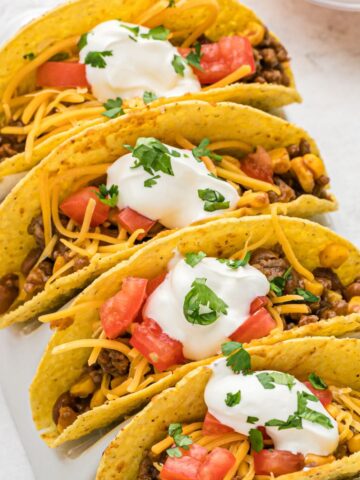 A row of tacos topped with sour cream, tomatoes, and filled with ground beef bbq mixture.