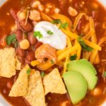 View of a white bowl with chicken chili in it and toppings like chips, sour cream, and avocado.