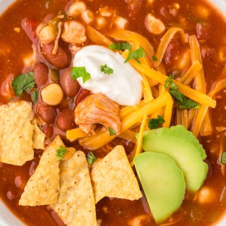 View of a white bowl with chicken chili in it and toppings like chips, sour cream, and avocado.