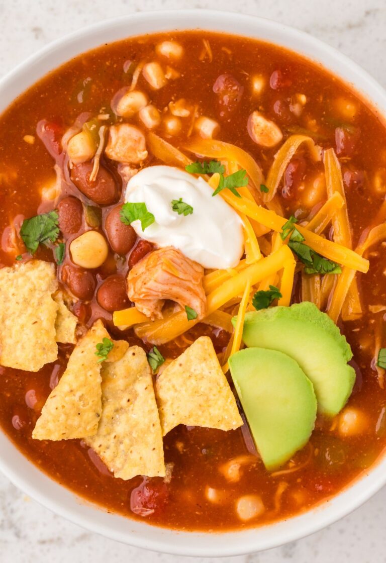 View of a white bowl with chicken chili in it and toppings like chips, sour cream, and avocado.