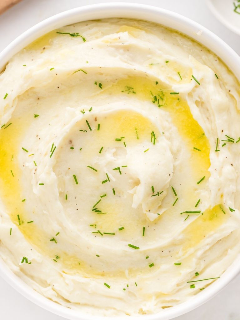 An overhead shot of a white bowl of mashed potatoes with melted butter and chives. 