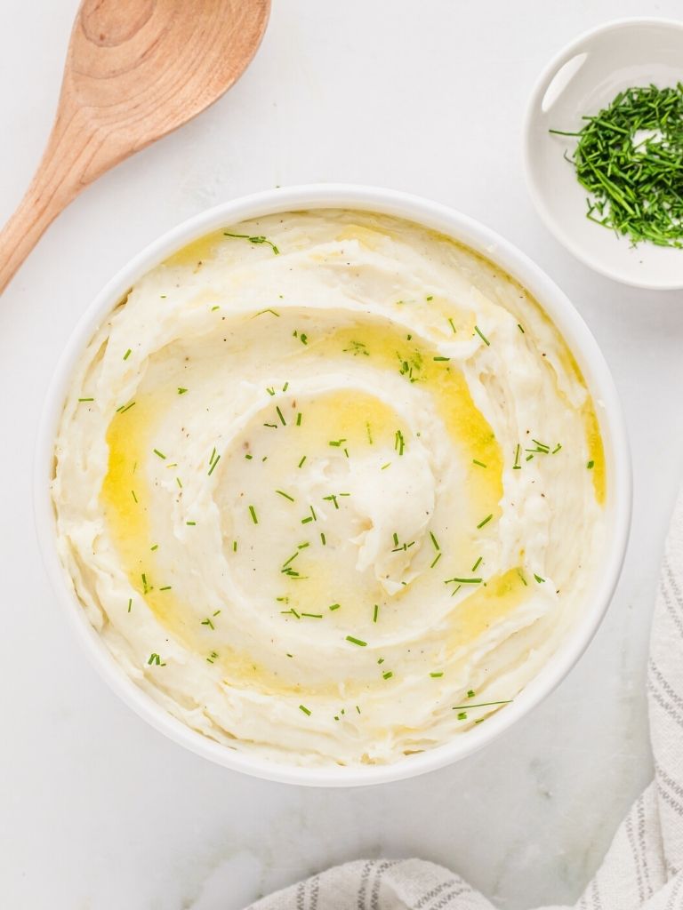 Overhead picture of a bowl of mashed potatoes with melted butter on top. Chives in a bowl in the background