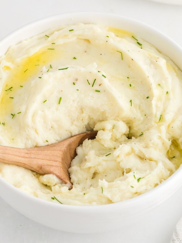 A dish with potatoes inside of it and a wooden spoon taking a scoop of them. 