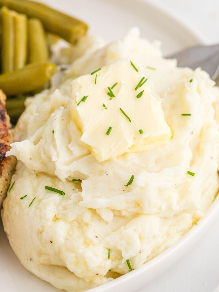A serving scoop of mashed potatoes with cream cheese on a white plate next to green beans. 