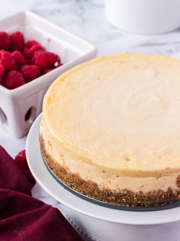 Cheesecake on a cake stand with raspberries in the background.