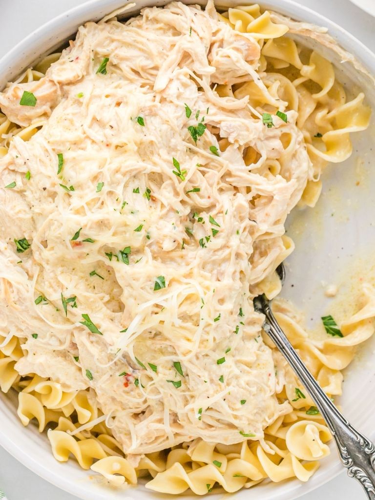 A white dish of chicken over some egg noodles with a serving spoon inside. 