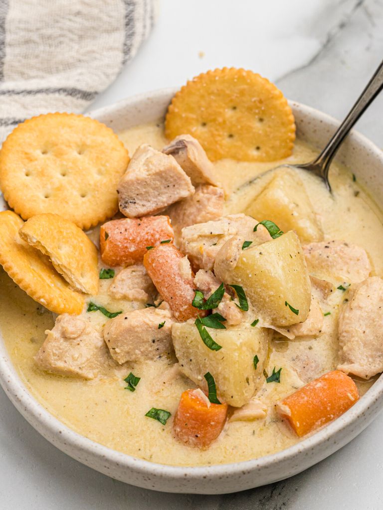 A bowl of stew with a spoon in it and some crackers to the side of it. 