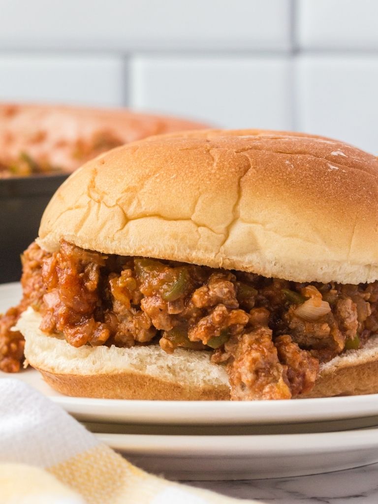 A sloppy Joe in a hamburger bun on a stacked pile of white plates. 