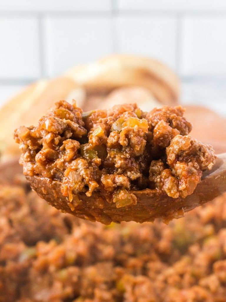 A wooden scoop of sloppy Joe mixture with a pan of it in the background. 