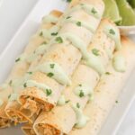 A stack of taquitos with sauce dripped over them and cilantro and lime slices in the background.