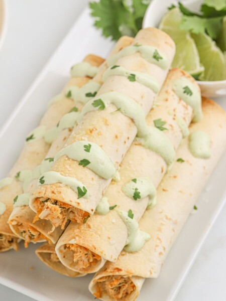 A stack of taquitos with sauce dripped over them and cilantro and lime slices in the background.