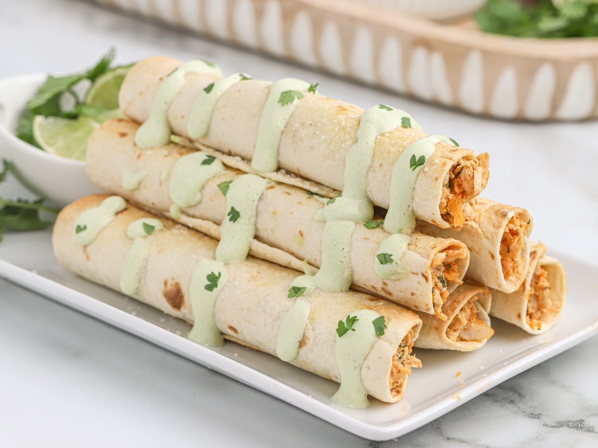 Image of taquitos on a white plate with salsa verde sauce dripped over them. 