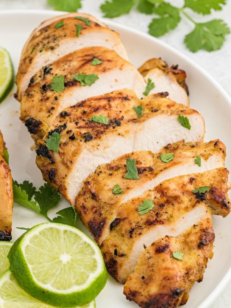 A grilled thai coconut chicken breast on a white plate cut into slices. Garnished with cilantro and lime wedges.