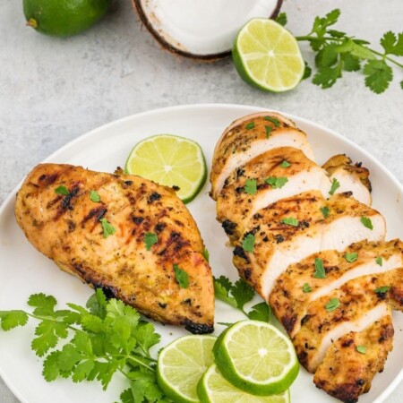 Grilled chicken on a white plate with one sliced and the other whole. Photo is garnished with lime, cilantro, and a halved coconut.