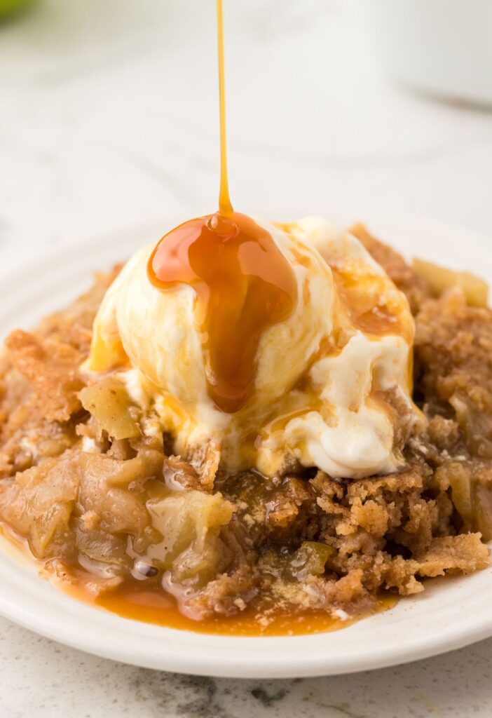 Serving of apple cobbler with ice cream and a caramel sauce being drizzled over top of it. 