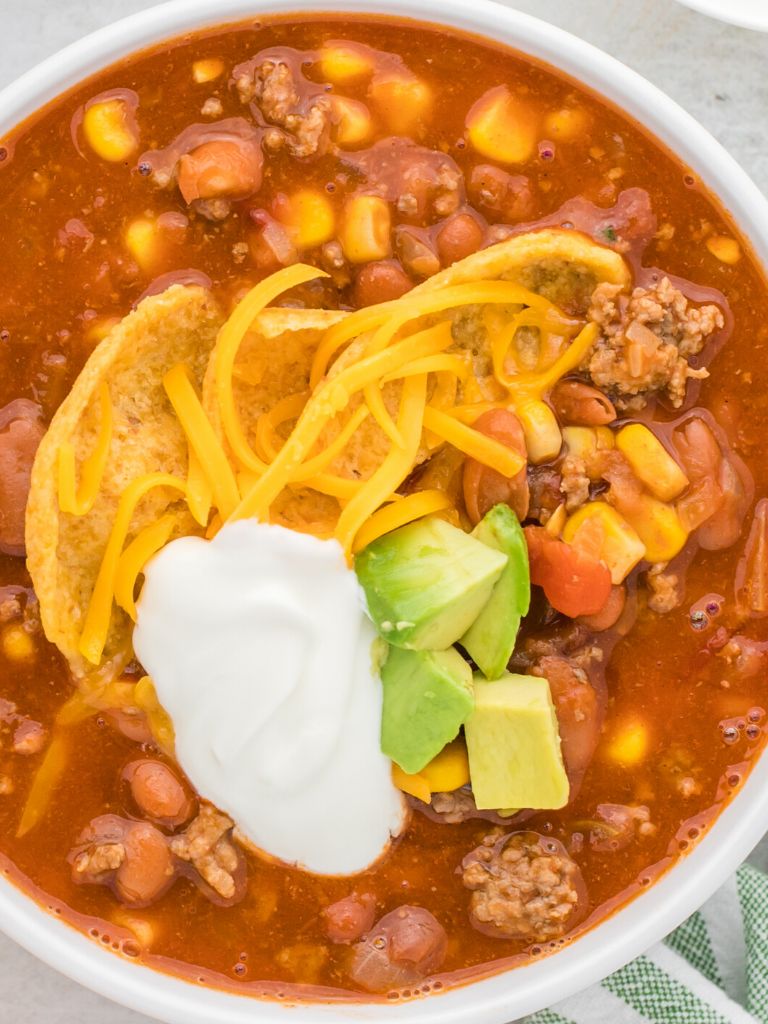 Overhead shot of a white bowl with taco soup inside of it and topped with sour cream, cheese, and avocado chunks. 