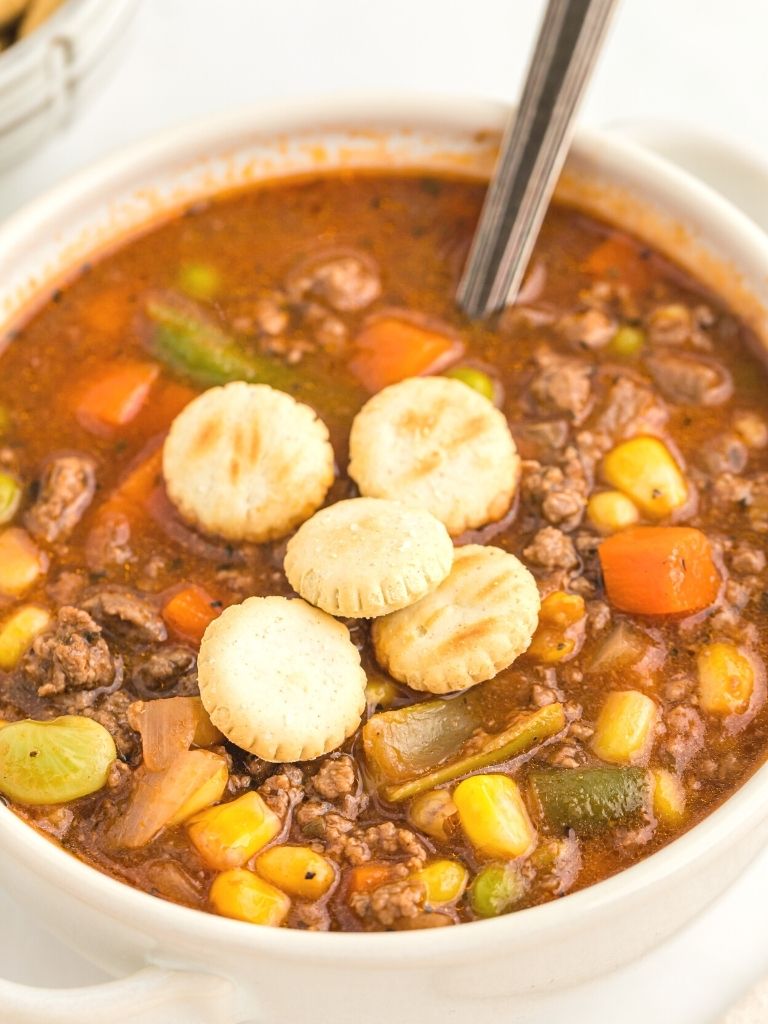 White bowl of vegetable soup with beef with oyster crackers on top of it and a silver spoon in the bowl.