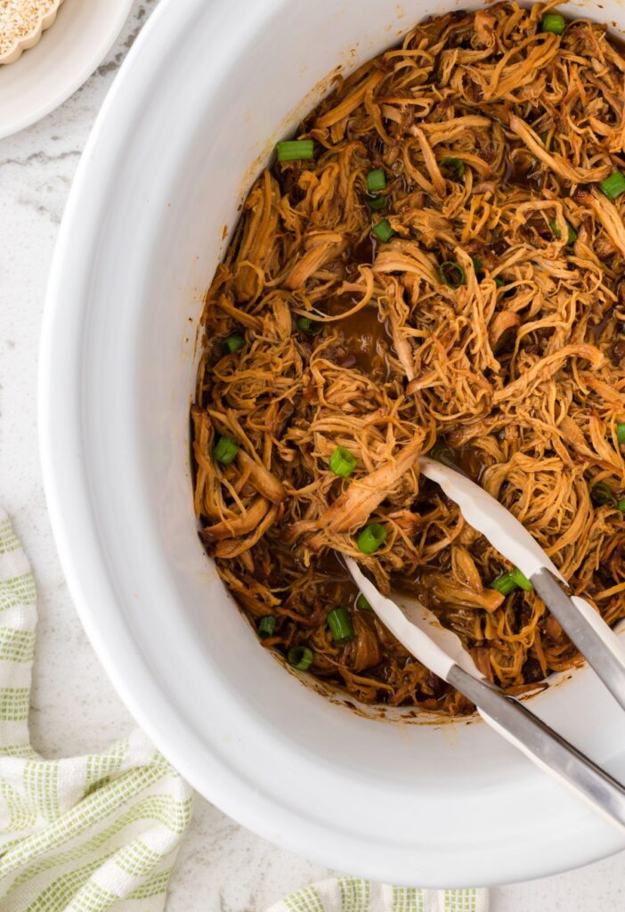 Inside of the slow cooker with tongs with some shredded chicken 