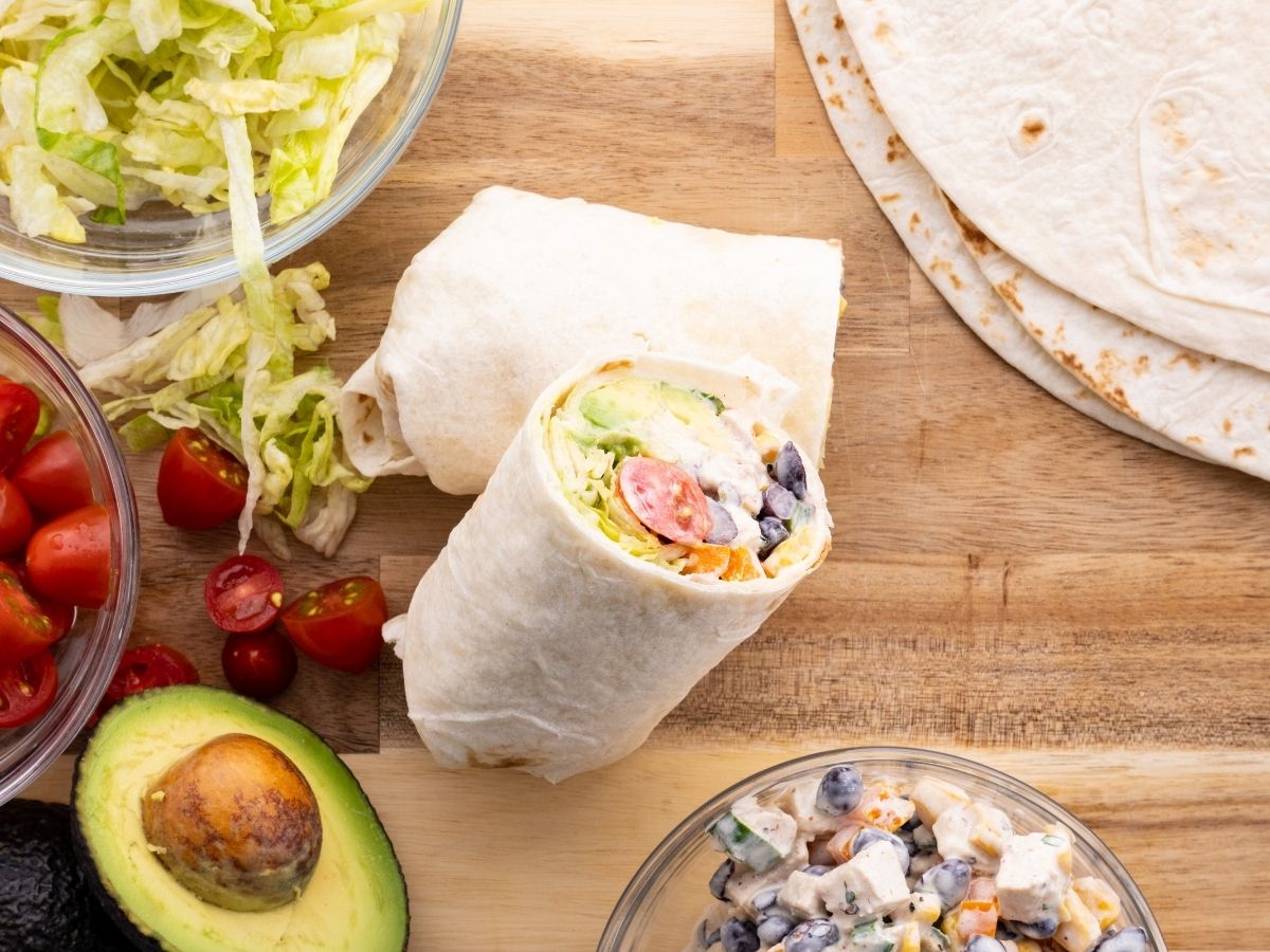 Horizontal photo of chicken salad, avocado halves, lettuce, and flour tortillas for wraps. 