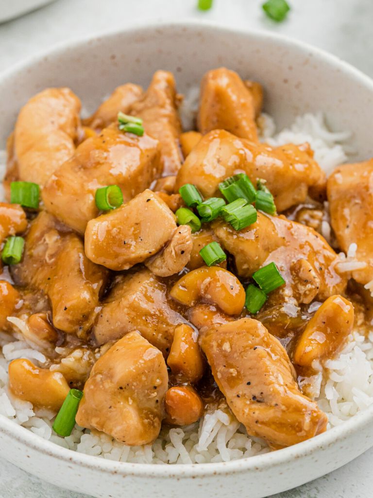 A white bowl of chicken with sauce and green onions, served over white rice. 