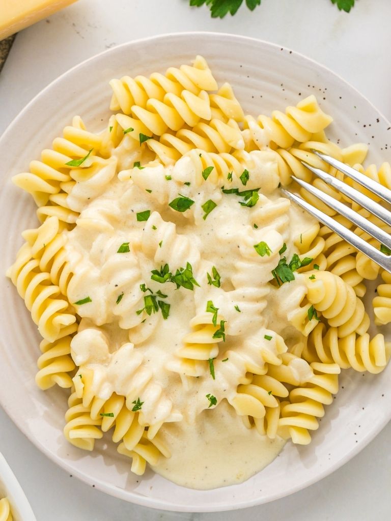 White plate with spiral pasta topped with alfredo sauce and parsley leaves. 
