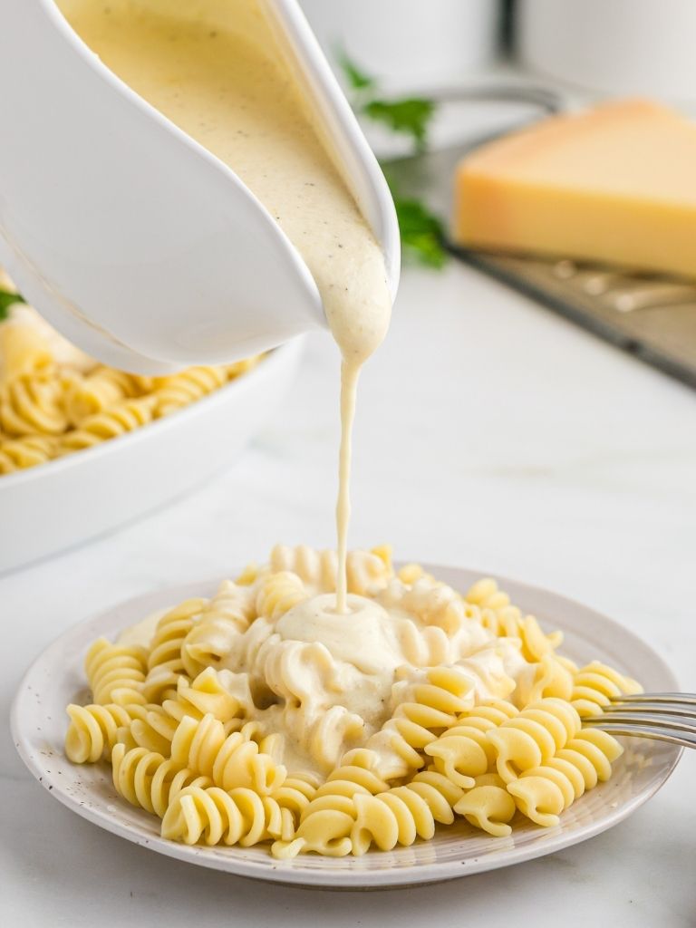 A gravy dish pouring alfredo sauce over cooked pasta on a white plate.