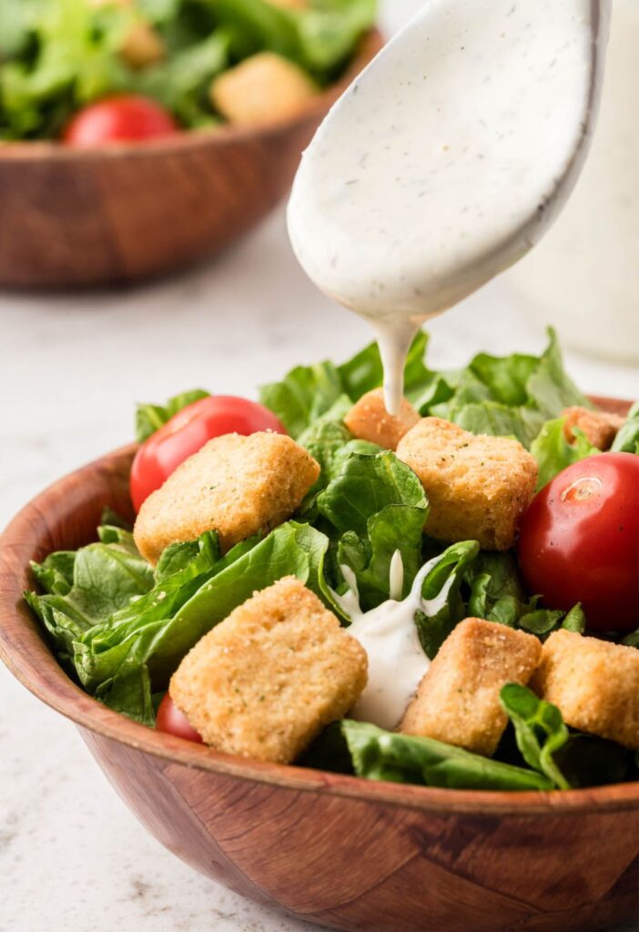 A bowl of green salad with ranch being poured over it from a silver spoon. 