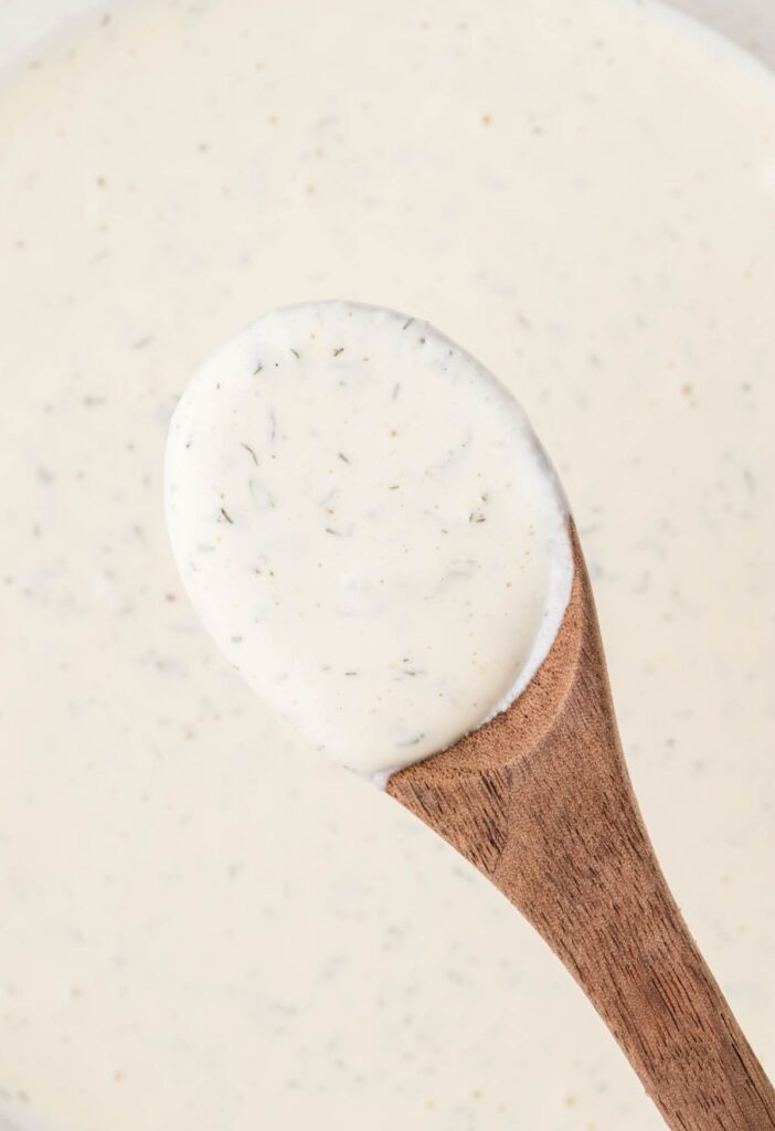 Close up of a bowl of creamy ranch dressing with a wooden spoon up close with ranch on it. 