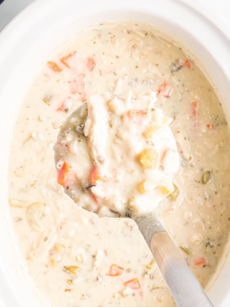 A slow cooker with creamy chicken wild rice soup with a scoop with some in it.
