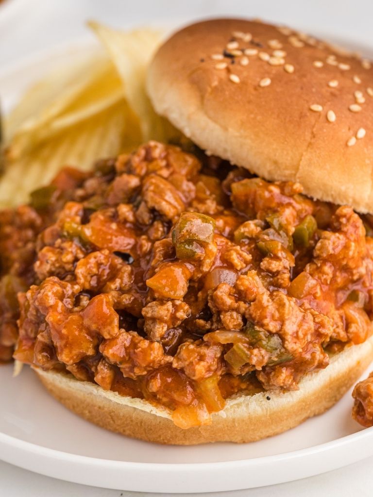 A white plate with a hamburger bun on it and chips in the background. Sloppy Joe meat on the bun. 