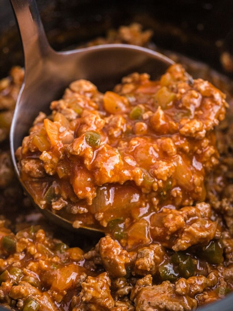 A close up picture of a silver serving spoon with sloppy Joe meat inside of it. 