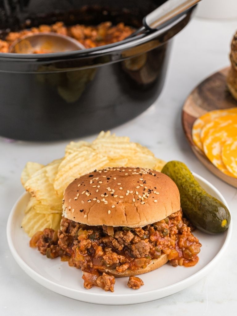 Slow cooker in the background with the meat inside of it and a white plate with a sloppy Joe sitting on it with a pickle to the side of the plate. 