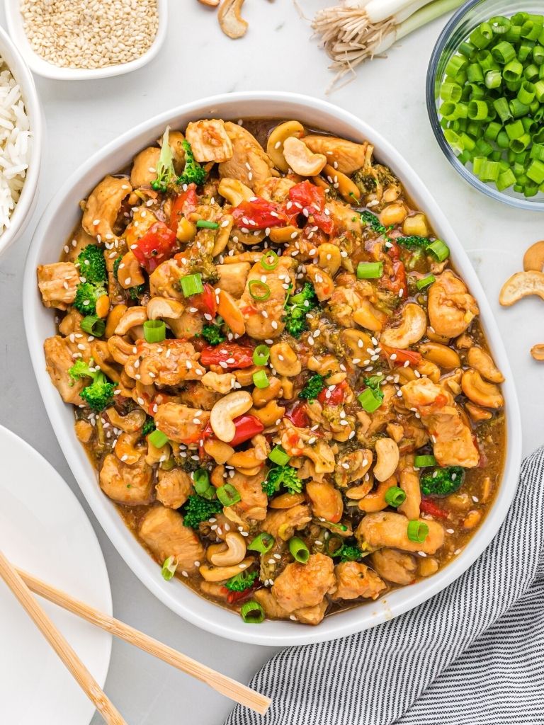 Overhead shot of cashew chicken on a white background with chopsticks, rice, and green onion on the side. 