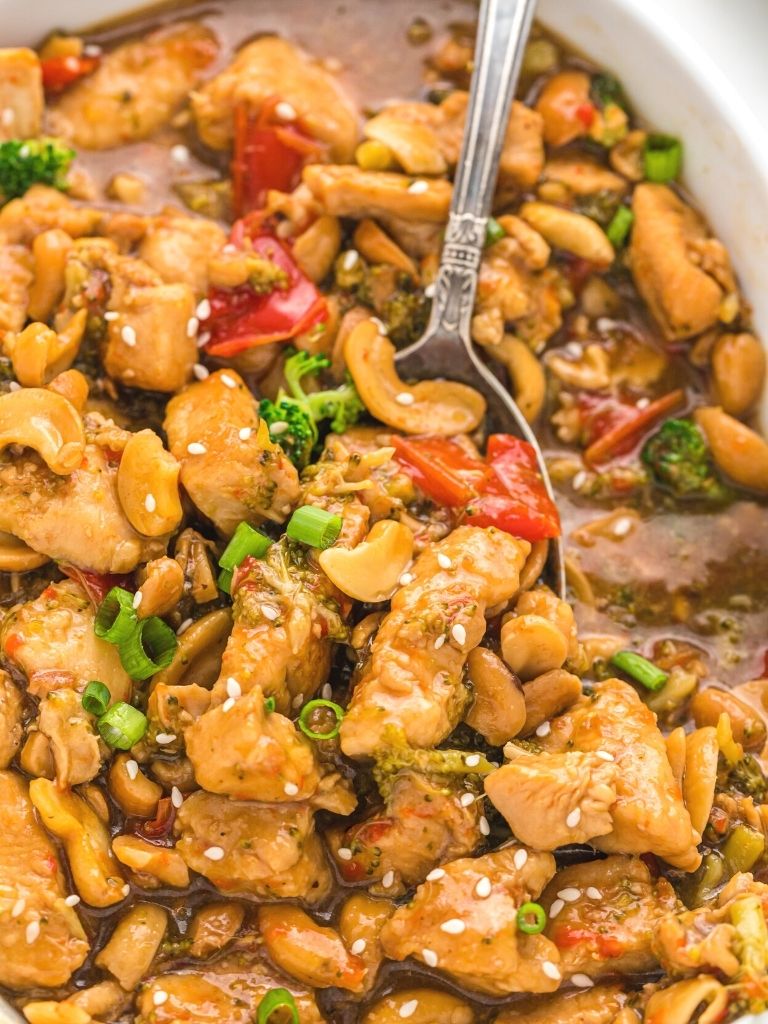 Close up shot of cashew chicken inside a white baking dish with a silver spoon in it. 